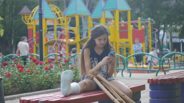 Girl with a broken leg sits on a bench, on the background of a descriptive pad and looks at the smartphone
