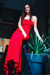 A slender girl in a red long dress is sitting on the railing on the steps near the house.Fashion photo