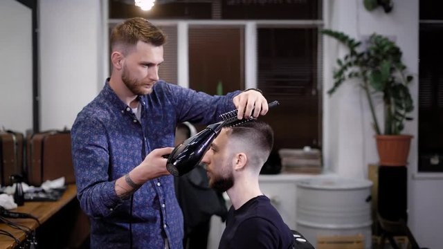 Male barber finishing haircut. He drying customers hair with hair dryer and comb. Barber shop scene