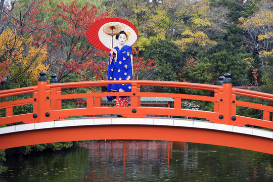 Asia, Japan, Honshu, Kansai Region, Kyoto, Geisha holding an ornate red paper umbrella standing on a traditional style Japanese red bridge 