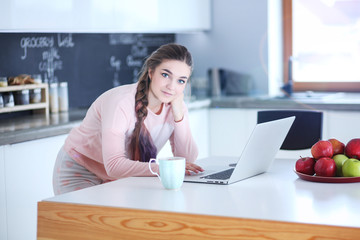 Young woman sits at the kitchen table using a laptop and talking on a cell phone.