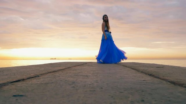 The young beautiful girl dances in a blue evening or ball dress on the pier close to sea. Slow motion.