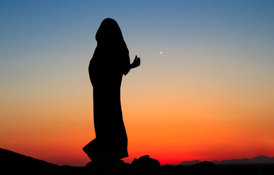 Arabian nights. Woman in black dress abaya with scarf around head and neck at sunset with mountains landscape and deep orange and blue sky, arabic mystic authentic style photo session in desert