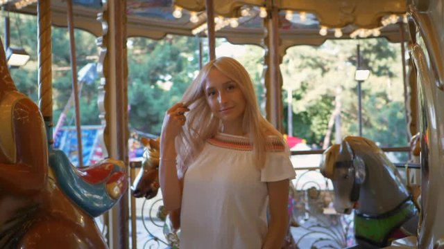 Close up of beautiful happy blonde woman having fun riding carousel in amusement park, smiling and waving at camera, graded. Slow motion.