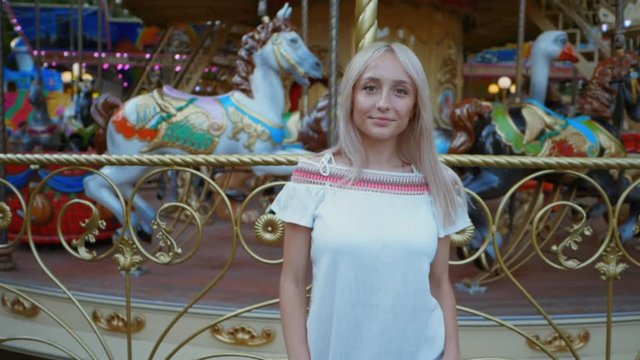 Young attractive blonde girl standing near carousel in amusement park. Beautiful portrait of woman with long hair. Slow motion.