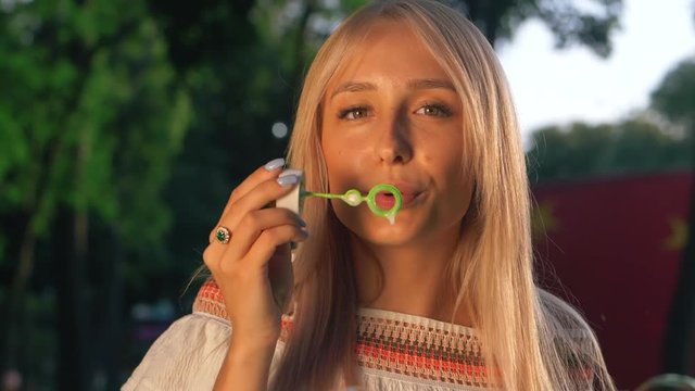 Beautiful young blonde girl blowing soap bubbles into the sky. Gorgeous young teen girl blowing bubbles. Outdoor summer portrait of attractive happy woman having fun in park or at nature. Slow motion.