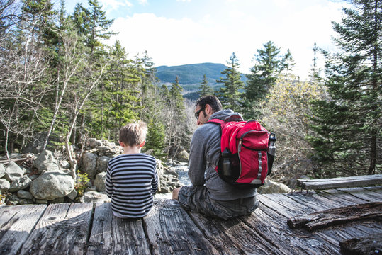 Boy and his dad rest together on a wooden bridge while hiking in the mountains