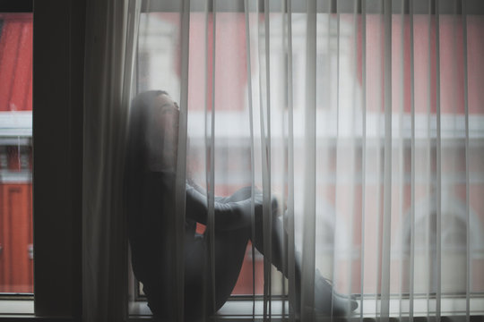 Woman sits at window behind curtains.