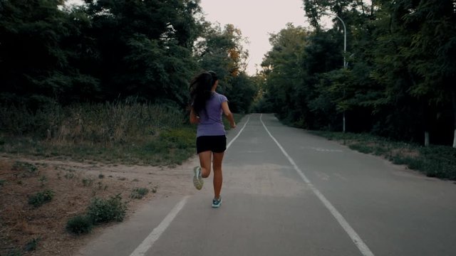 Girl runs through the park in the morning. Sporty young attractive brunette woman running on empty road in the morning. Slow motion. Back view.