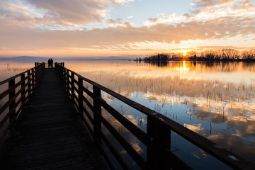 A first person view of a pier on a lake with two people on it, beautiful sunset and clouds reflecting on water
