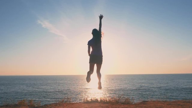 Woman jumping with excitement, slow motion. Silhouette of Young Active Sporty Successful Woman Standing on the high hill above sea or ocean and enjoy sunrise.