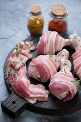 Black wooden chopping board with raw fresh chicken legs wrapped in bacon, close-up, studio shot