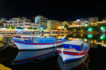 Fototapeta na wymiar Fishing boats at para outra in night city of Crete, Agios Nikolaos in the background of seaside restaurants, cafes and hotels. Tourists relax in outdoor restaurant on the shores of lake Voulismeni