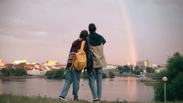 Back view of young hipster couple hugging on the shore of the river and taking photos of rainbow on smartphone.