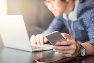 Young Asian businessman dressed in casual style holding smart phone and working with laptop computer in coffee shop. Freelance job and IT modern lifestyle, work life balance concept
