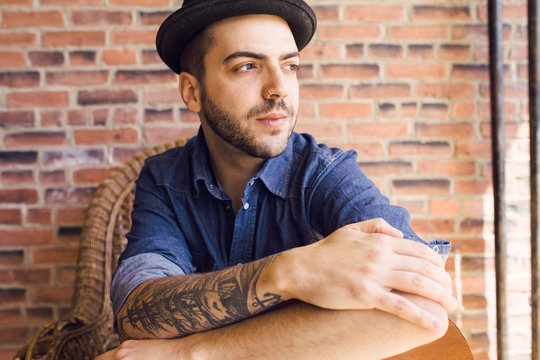 young tattooed brazillian man wearing a fedora looking out window