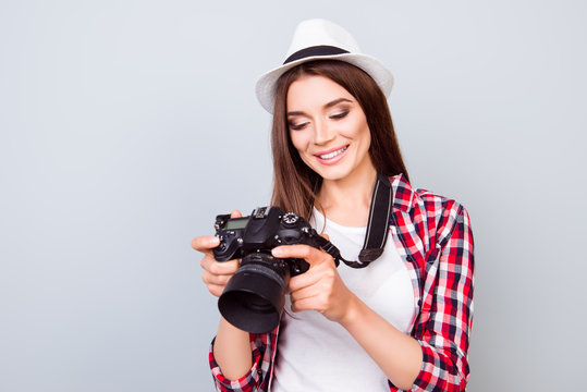 Young attractive brunette photographer is smiling on the blue background. She is excited and holding camera, wearing summer  hat, watching the photos