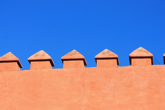 Minimalist architectural Spanish detail showing a warm terracotta-colored fortress or historic wall with evenly spaced decorative battlements against blue sky. Strong geometric composition 