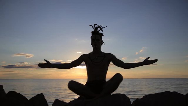 Silhouette of a man with native american indian feather mohawk accessory on head practicing yoga at the sunset.