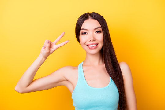 Peace! Young cute asian girl, standing on the bright yellow background and smiling, wearing blue casual singlet and showing vsign