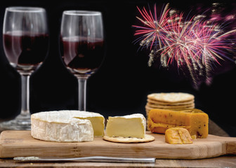 Brie and hot pepper jack cheese on wooden cheese board, accompanied by crackers and two glasses of red wine, against black background decorated with colorful fireworks