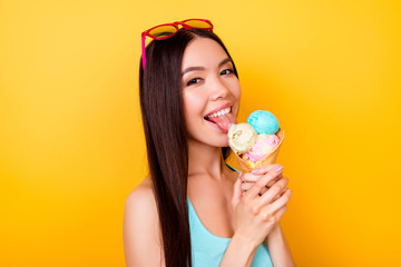 Excited young asian lady licks tasty ice cream of three scoops of different flavors, stands on yellow background in tourist wear, glasses