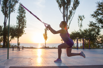 Fototapeta na wymiar Beautiful fit woman in pink and purple sportwear training on outdoor gym in morning, exercises with suspension straps in park