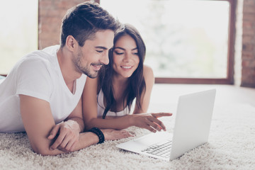 Beautiful latino mulatto lovers are browsing info on laptop and discussing it, lying on the floor on comfortable carpet at home. They are smiling, so relaxed