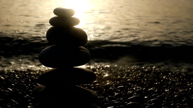 Closeup of pyramid made by sea stones on stony seashore at blurred golden light sea water background. Beautiful nature landscape with splashing waves of dark water. Real time video footage