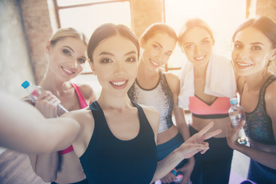 Selfie time, girls! Five girlfriends in fashionable sport outfits are posing for a selfie photo, that asian brunette is taking. They are all smiling, joyful after the workout