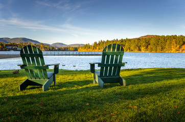 Fototapeta premium Two Green Adirondack Chairs on the Shore of a Mountain Lake at Sunset. Lake Placid, NY