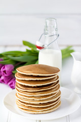 Simple Stack of Pancakes on the White Wooden Table with Bottle of Milk and Purple Tulips on the Background
