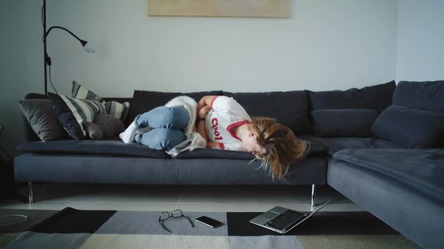 Poor young girl suffers from pain in her belly lying on big sofa at home. Blonde female teenager has strong ache because of her period, spinning on sofa and hopelessly looking advises in internet