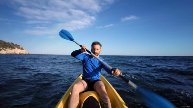 Strong and skilled man paddles on kayak in sea, portrait shot. Blue wavy ocean waters near shore with handsome bearded sporty man is kayaking forward, wearing lycra and shorts