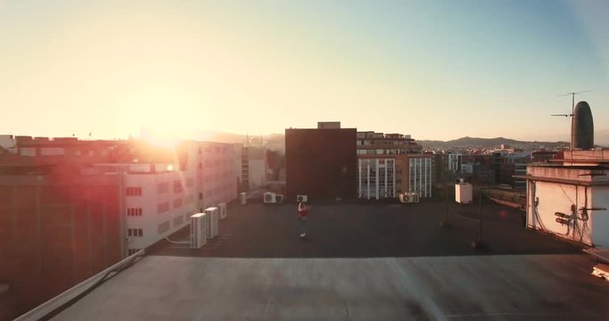 Aerial, scenic and dramatic shot of tiny small teenager girl is skating on her longboard on rooftop at sunset, drone flies toward and away to show industrial gentified district at summer evening