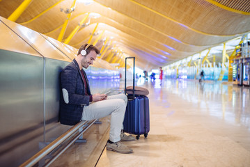 Young man waiting listening music and using mobile phone at the airport with a suitcase.