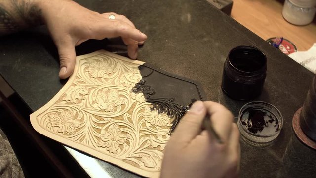 An adult bearded skinner paints a black piece of leather with amazing embossed flowers close-up 4k.