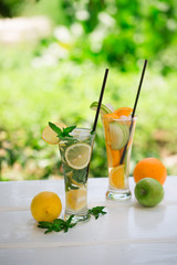 Two cocktail glasses of non-alcoholic mojito and detox fruit water orange-green apple and lemon-mint leaves on  white wooden table outdoors with garden background