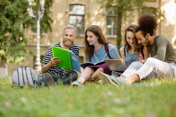 Fototapeta premium View on students sitting on lawn studying in college garden. College campus, young men doing teamwork together.