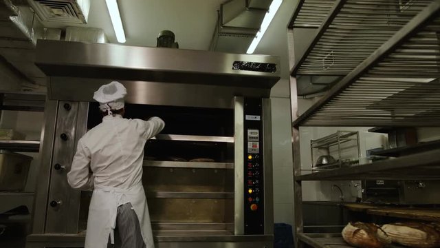 Young man in the uniform removing breads from oven and putting it to the reck. View from the bottom.