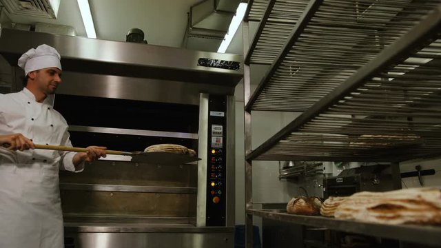 Baker man in the uniform taking breads from oven and putting it to the reck with other breads.