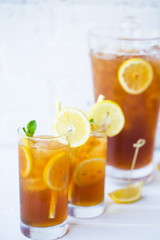 Two Glasses and Pitcher Filled with Iced Tea Ice Cubes and Lemon Slices Garnished with Mint and Striped Straws Against the White Brick Wall on the Wooden Table Close UP