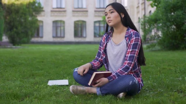 Thoughtful Asian female student sitting on lawn alone and missing parents