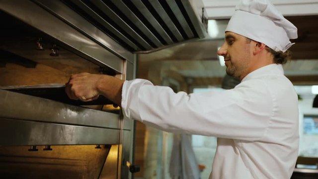 Portrait of young man in the uniform putting rolls to the oven in the modern bakery. Close up shot.