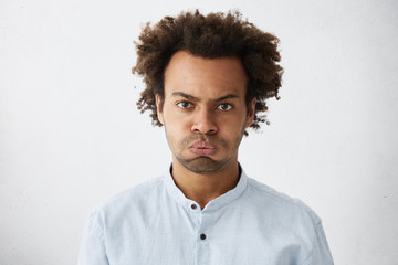 Fototapeta premium Isolated studio shot of unhappy grumpy serious young African American manager with Afro hairstyle frowning, puffing his cheeks, feeling fed up as he has to stay at office and work extra hours.
