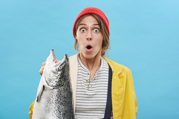 Attractive amazed female wearing red hat and yellow anorak holding huge fish in hand having shock to catch it. Young female expressing surprisment looking with bugged eyes while standing with fish
