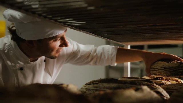 Close up shot of young baker standing in his bakery in the morning and sorting the fresh brown bread from the rack. Close up shot.