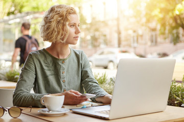 Portrait of blonde female with stylish hairstyle dressed formally sitting in front of opened laptop using cell phone resting at restaurant drinking hot coffee. Successful businesswoman working online