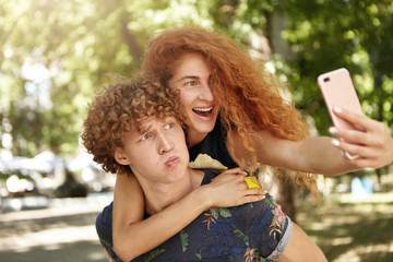 Outdoor portrait of curly male holding on back his girlfriend with freckled face and red hair looking at camera of cell phone, making selfie together. Positive redhead female and her boyfriend in park