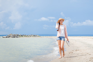 Fototapeta na wymiar A young woman in a white T-shirt, denim shorts and a pink knitted cardigan holds a straw hat and walks next to the sea on a summer day